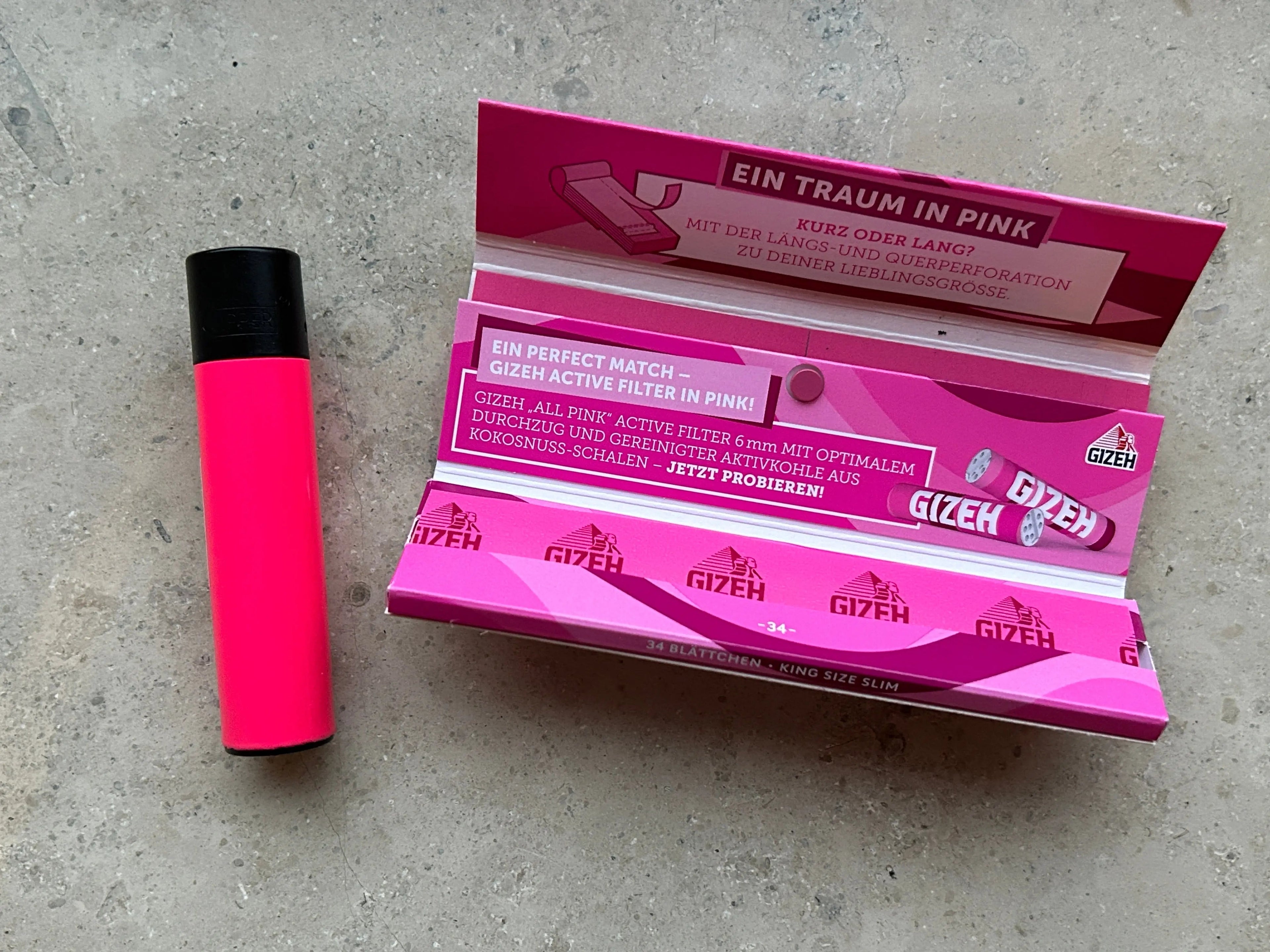 Ein rosa Clipper-Feuerzeug liegt neben einem offenen Ladys Starterpack von  das rosa Gizeh-Zigarettenfilter enthält, auf einer Marmoroberfläche. Das Paket ist mit mehreren Spitzen gefüllt und zeigt einen deutschen Text, der für das Produkt wirbt.
